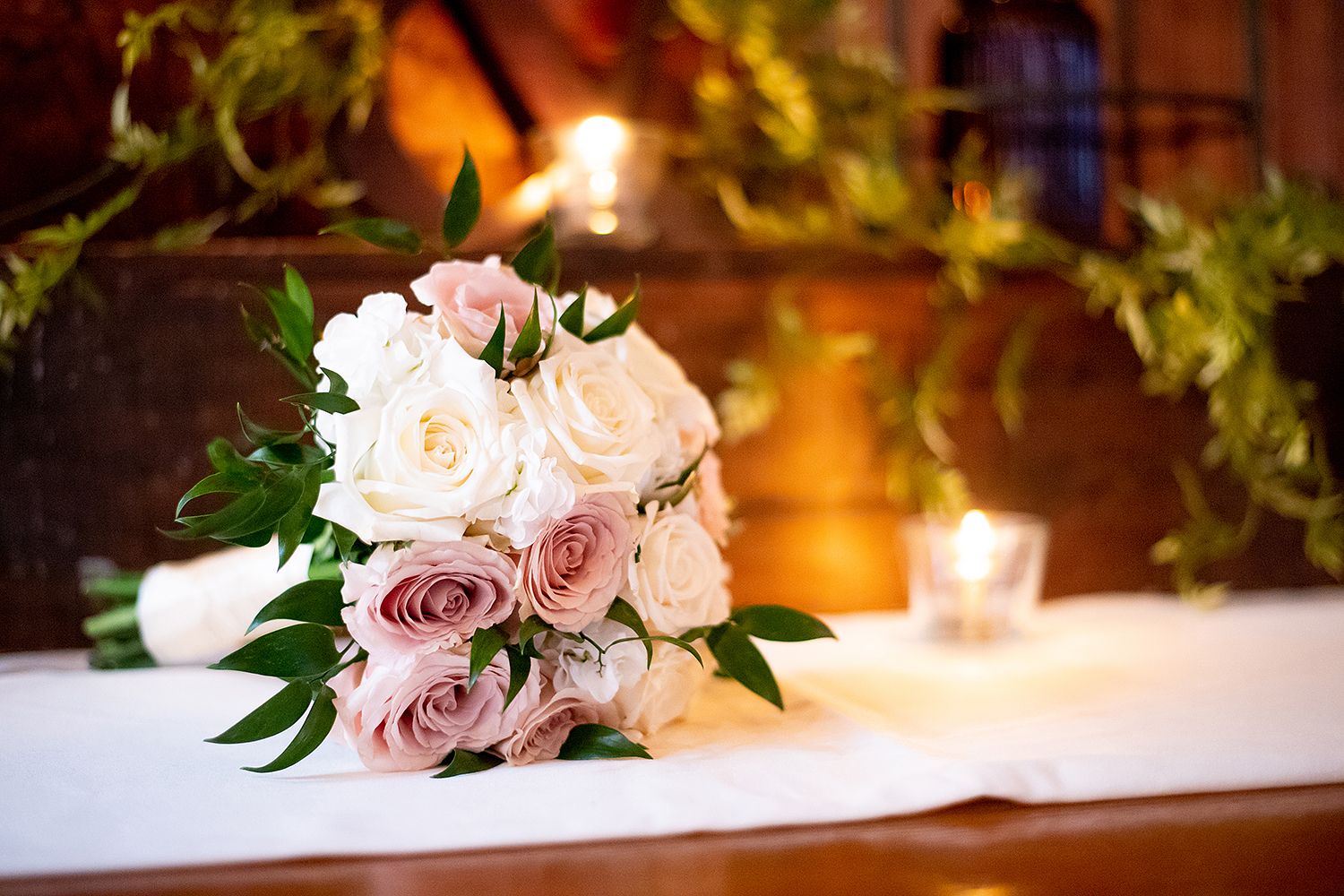 A blush pink rose bridal bouquet sits on white linen with candlelight and greenery in the background.