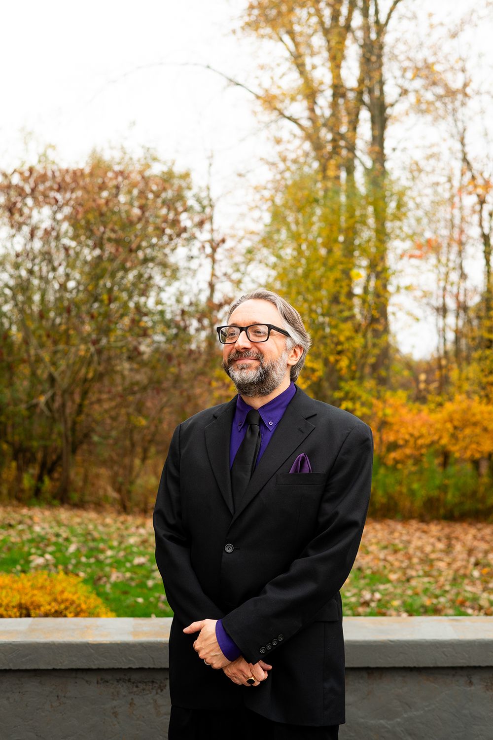A smartly dressed person in a black suit and purple tie stands outdoors against a backdrop of autumn foliage.