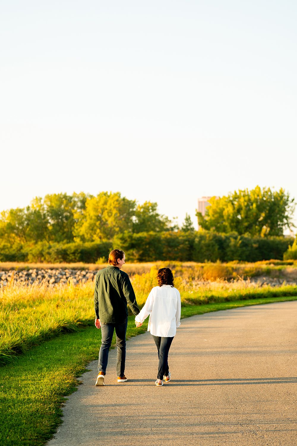 Two people walk together at sunset on a peaceful countryside path bordered by golden fields and green trees.
