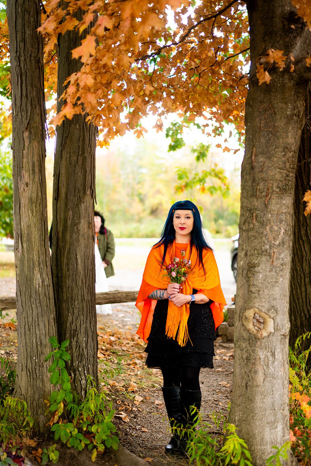 A figure in a bright orange poncho stands on an autumn forest path holding a bouquet beneath orange leaves.