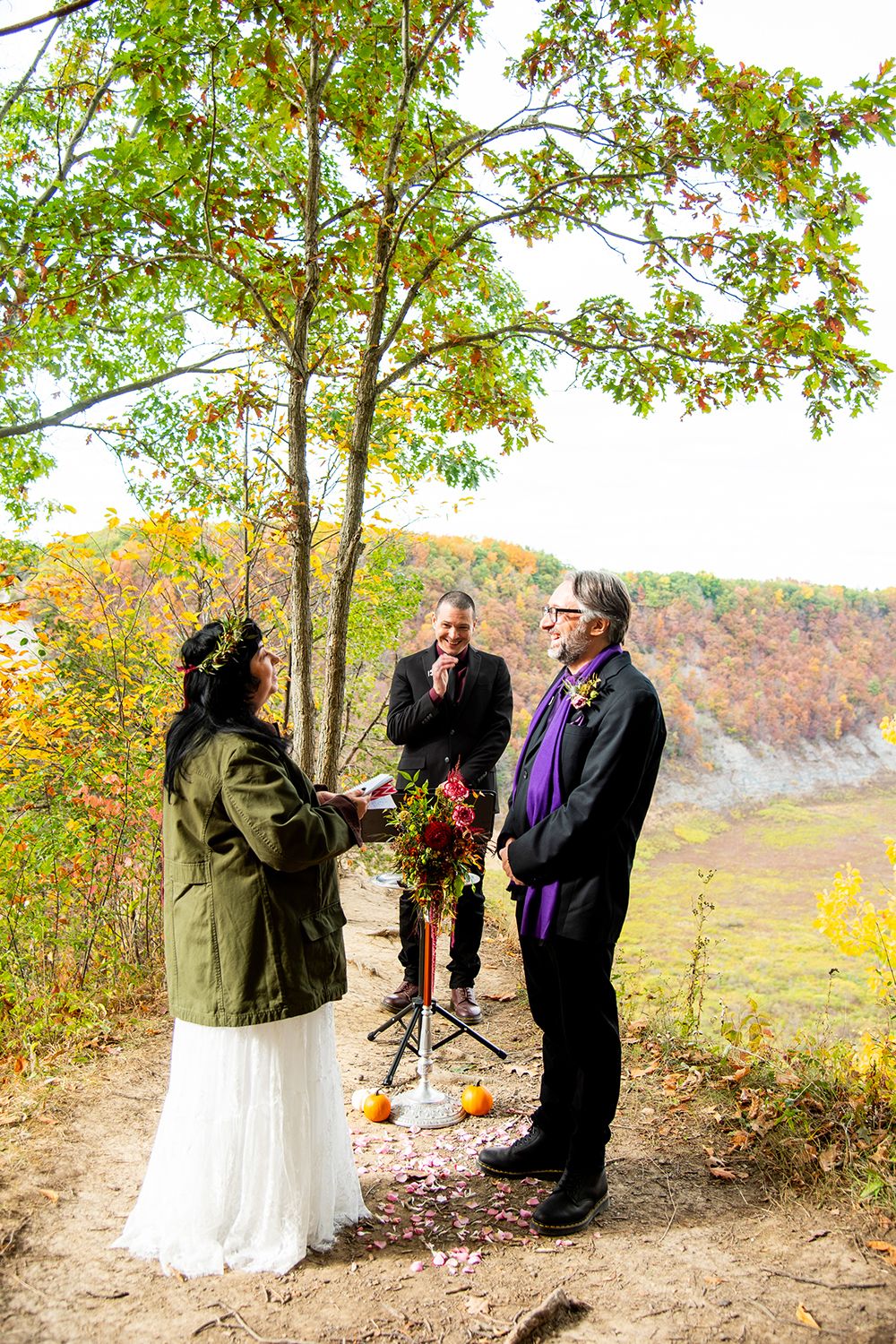 A fall wedding ceremony takes place on a scenic trail with autumn foliage and small pumpkin decorations.