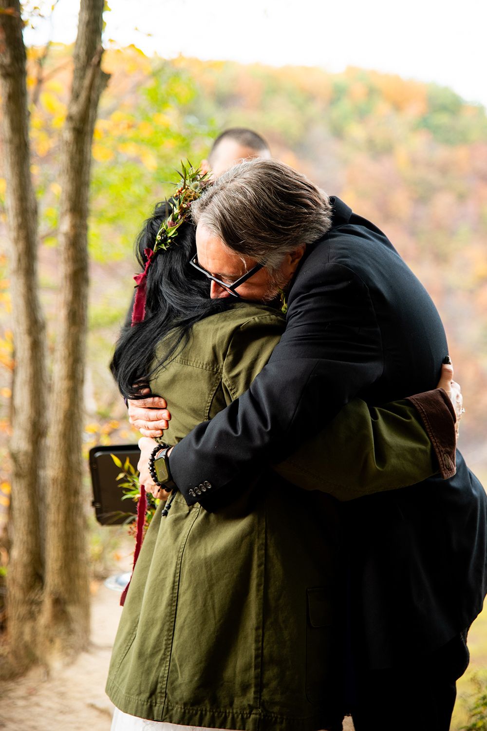 Two people share an emotional embrace on an autumn hiking trail surrounded by trees with fall foliage.