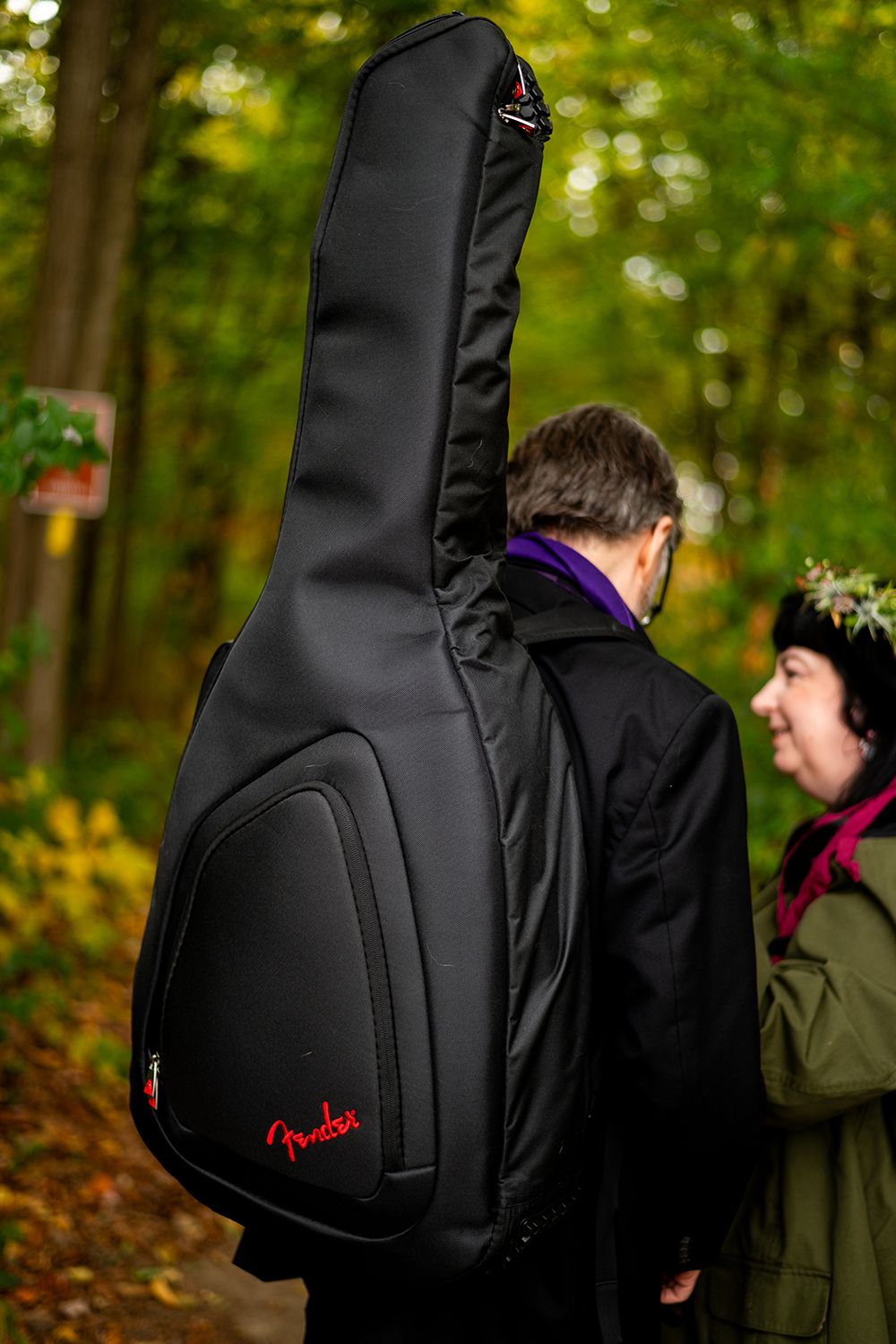 Black Fender guitar case with red logo is carried on someone's back in an autumn forest setting.