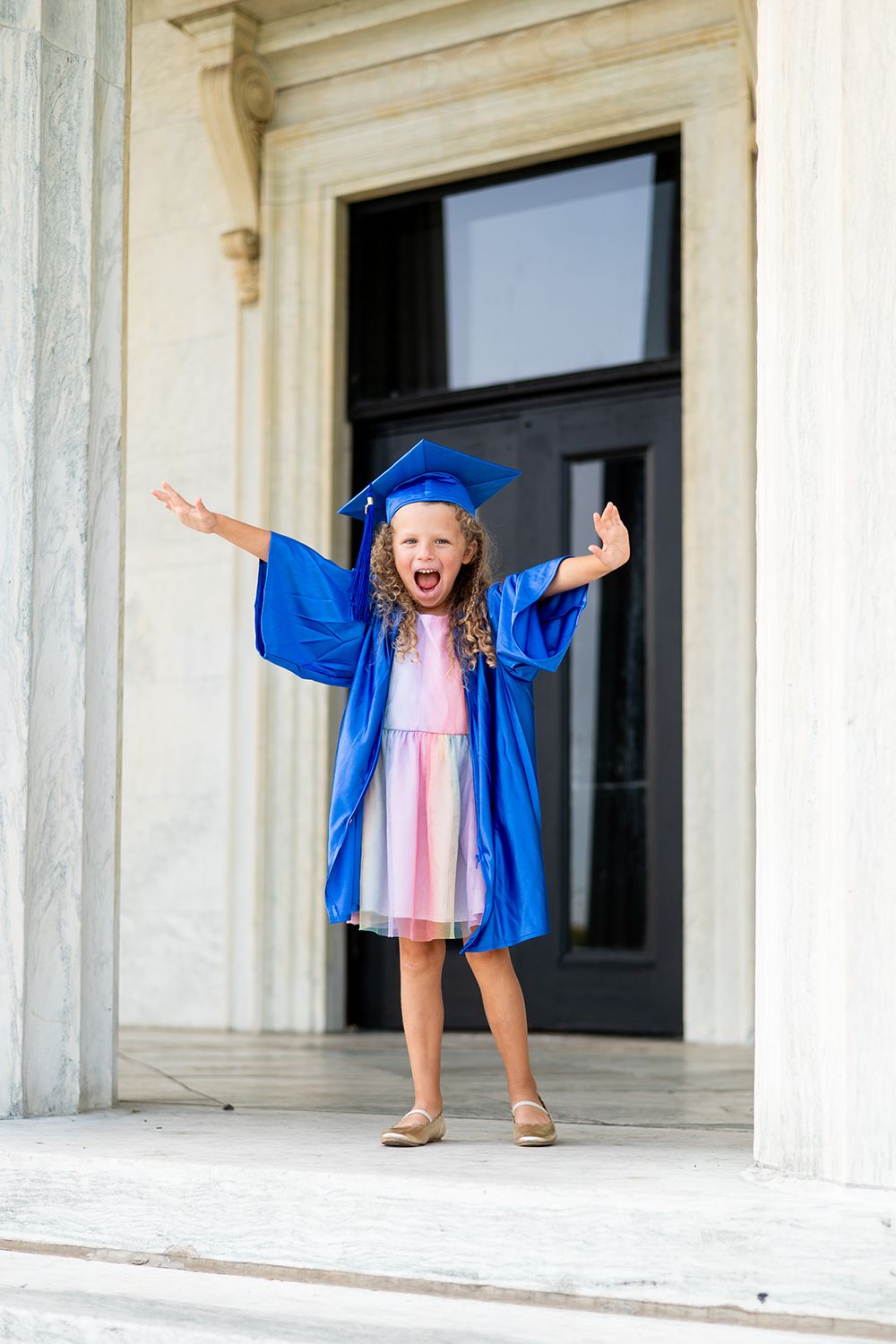 Graduate in bright blue cap and gown shows excitement with outstretched arms while standing in front of columned entrance.