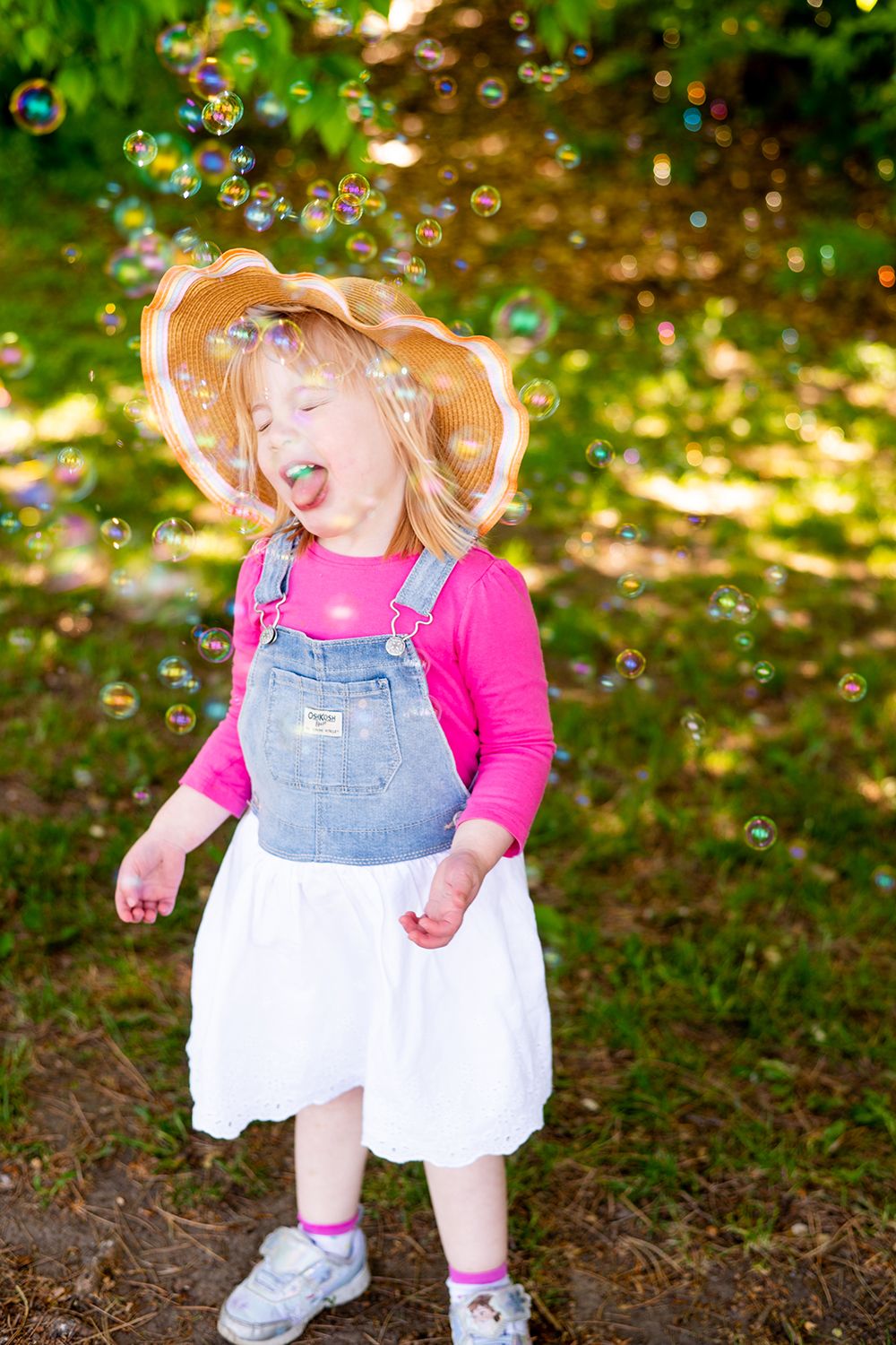 A young child in overalls and a sun hat plays joyfully among floating soap bubbles in a sunlit garden.