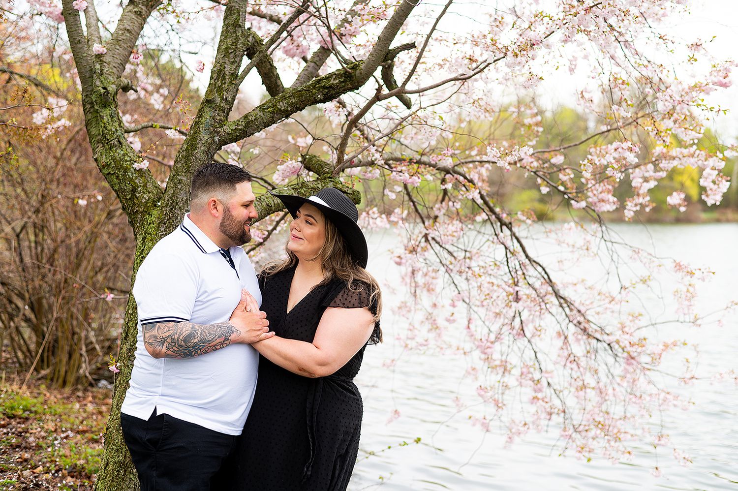 A couple embraces under blooming cherry blossoms by a tranquil lake in springtime.
