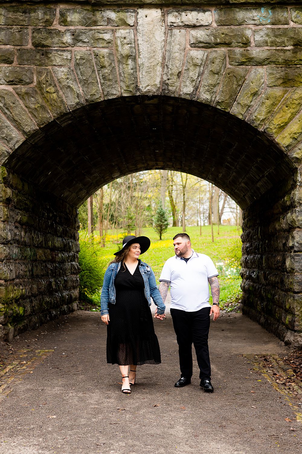 A couple walks hand in hand through a stone archway tunnel leading to a green spring forest path.