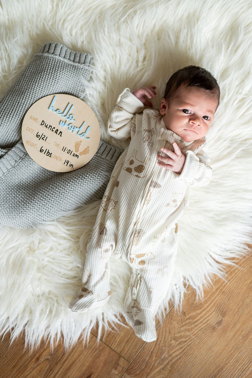 Baby lying on white fluffy blanket next to wooden milestone marker wearing cream colored sleeper.