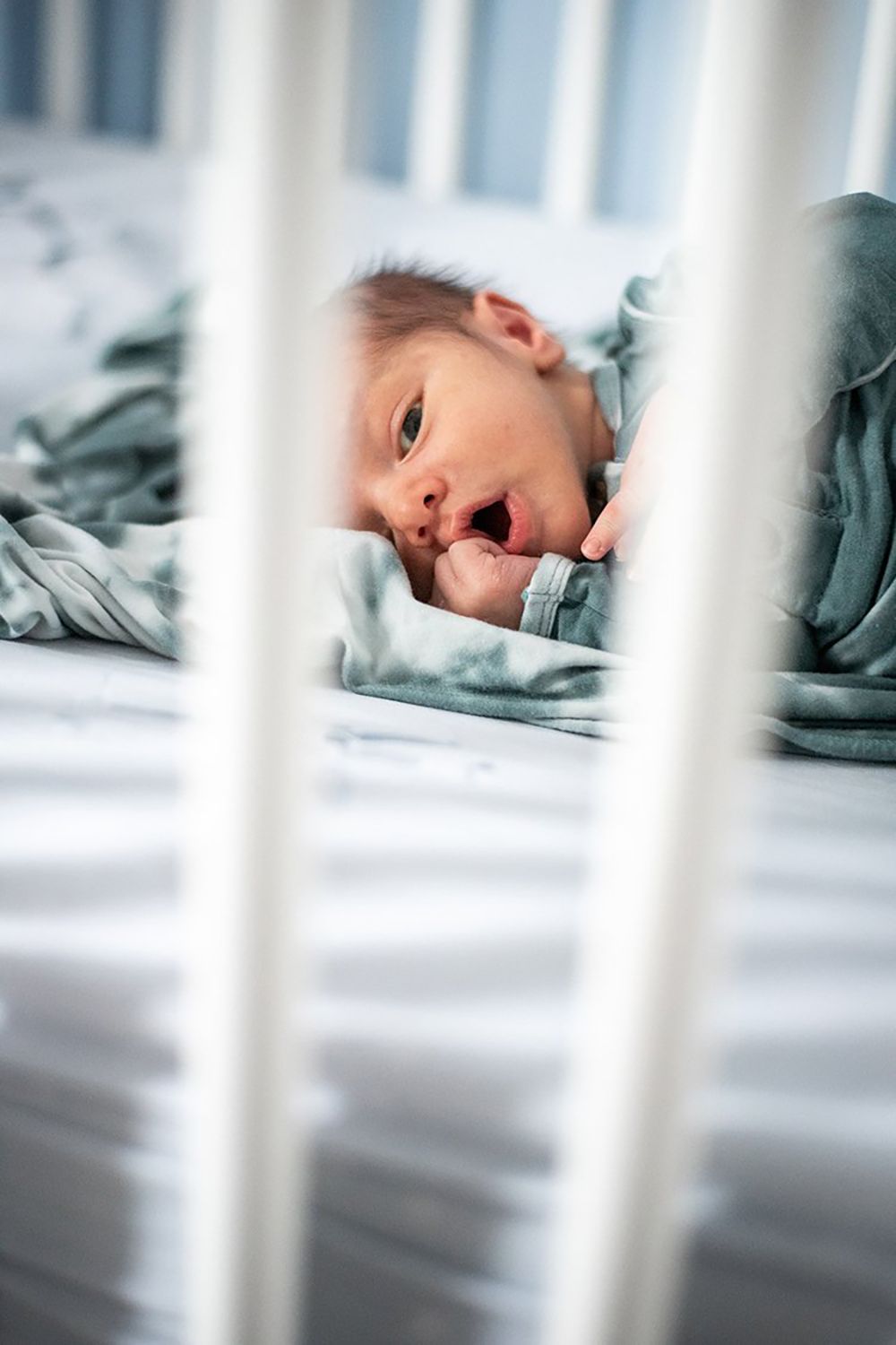 Baby lying in crib with white bars, wrapped in soft teal blanket while yawning or making facial expression.