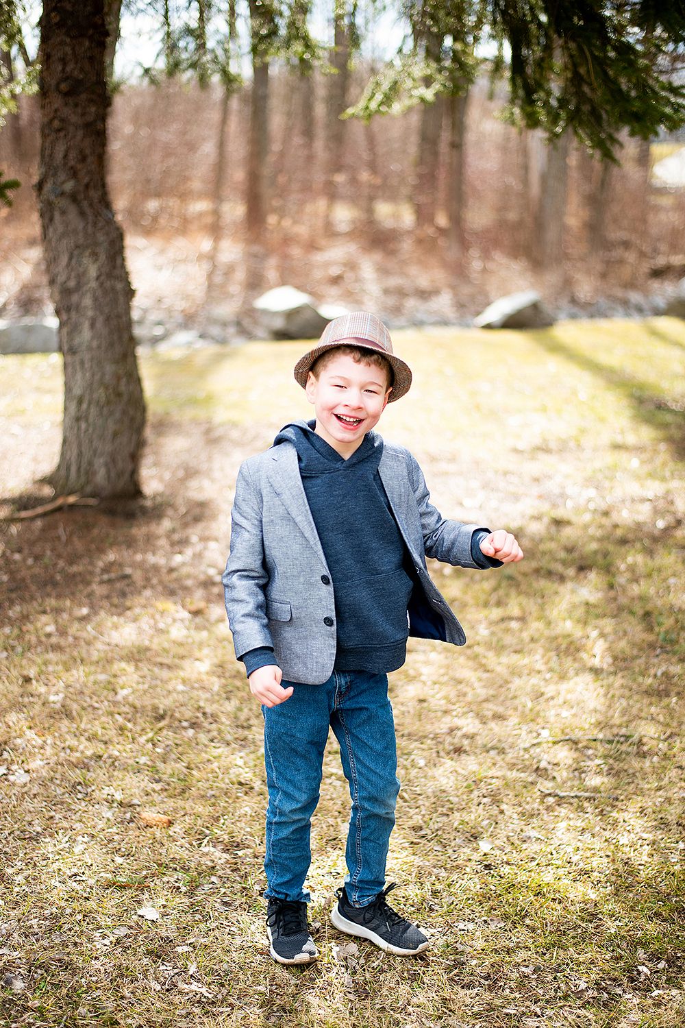 Young child wearing a beige hat, blue blazer and jeans poses playfully outdoors under a tree on grassy ground.