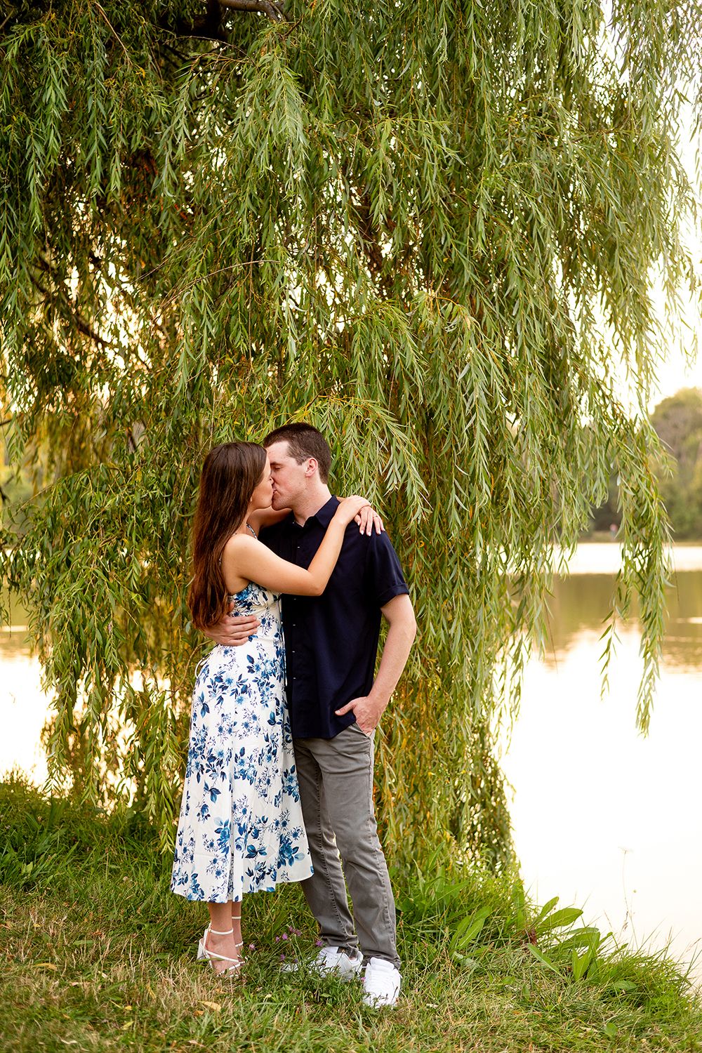 A romantic moment unfolds beneath drooping willow branches near a serene lake.