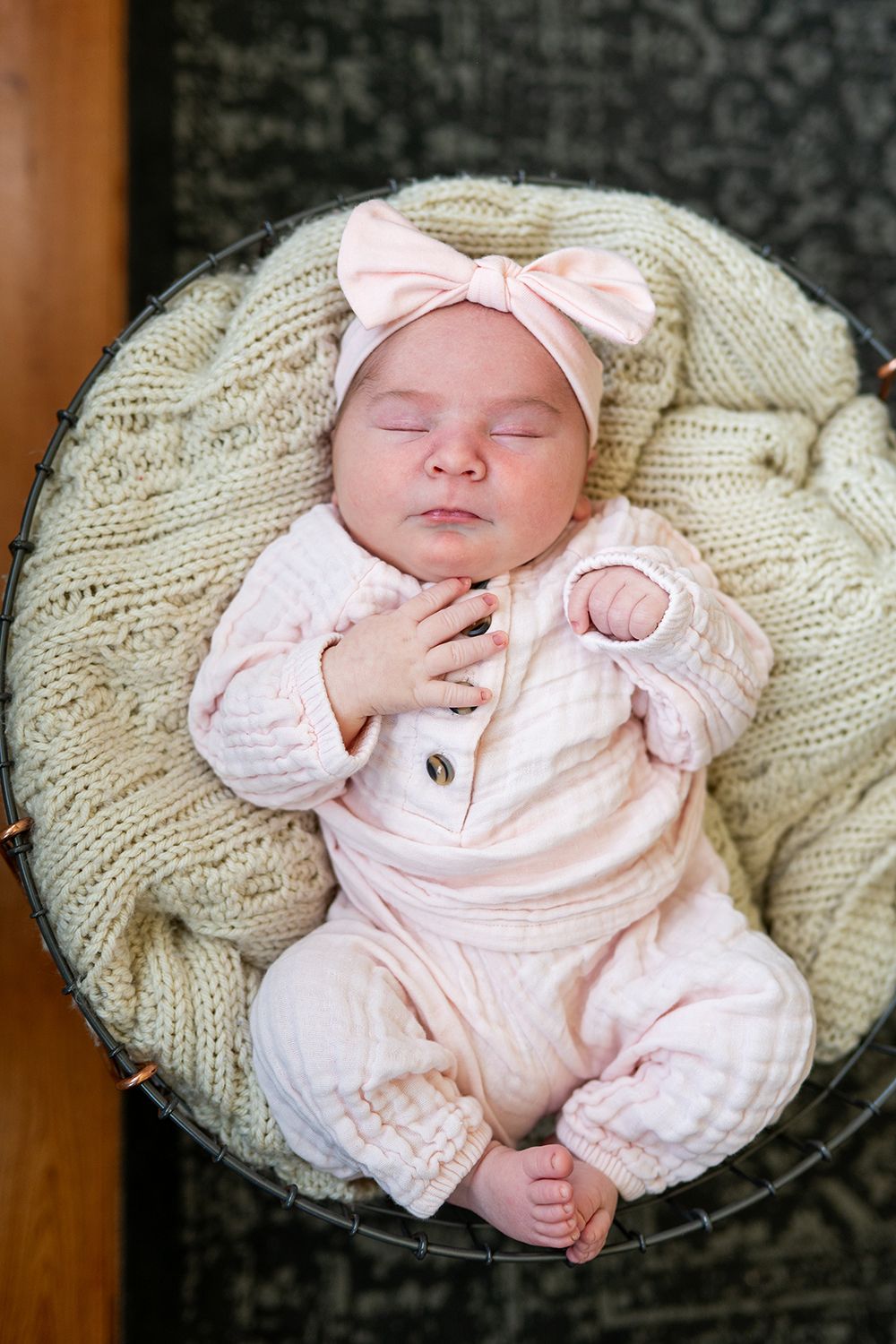 A sleeping baby wearing pink clothing and bow rests on a chunky knit cream blanket.