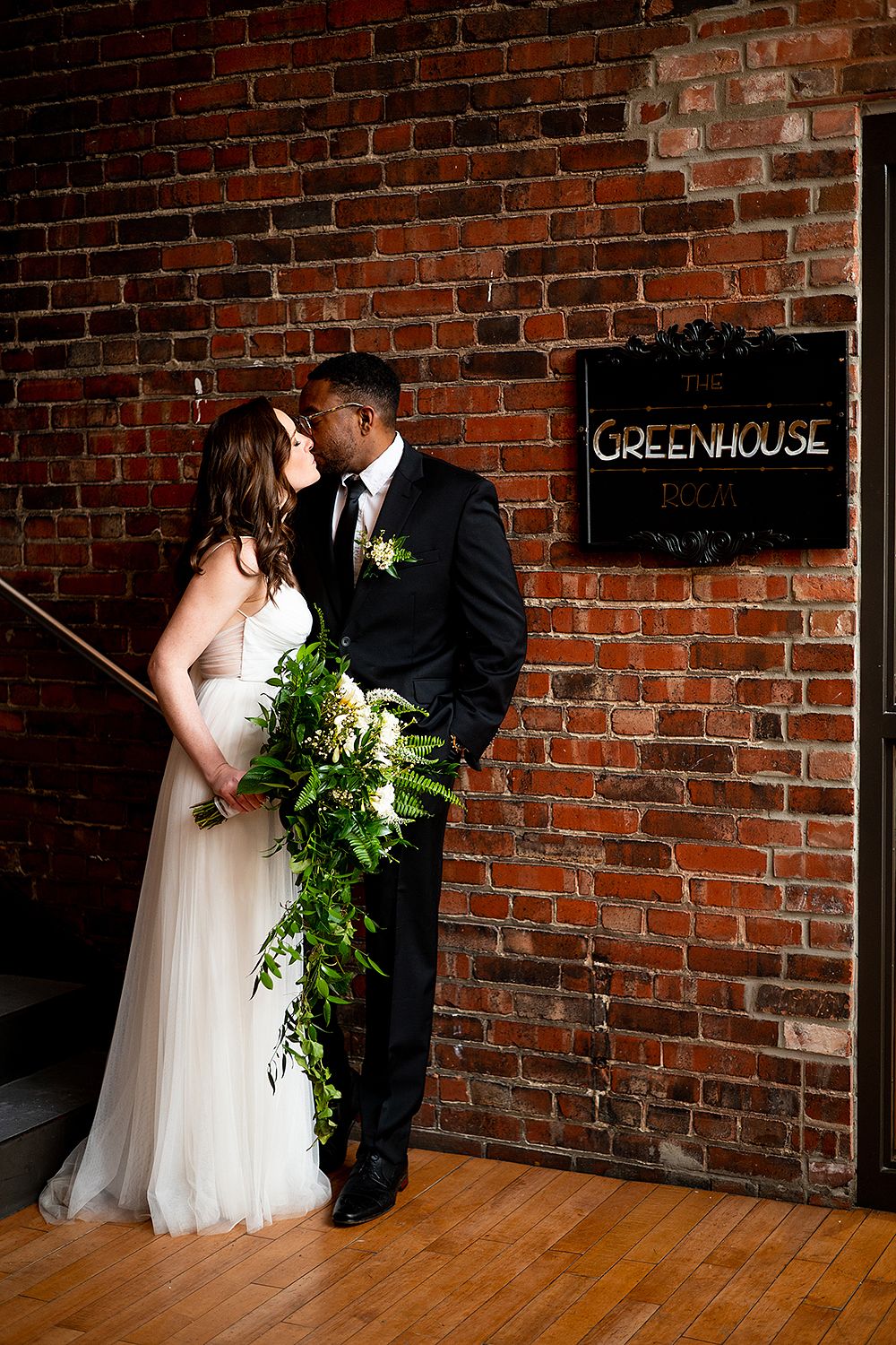 Couple poses intimately against rustic brick wall with greenhouse sign in elegant wedding attire.