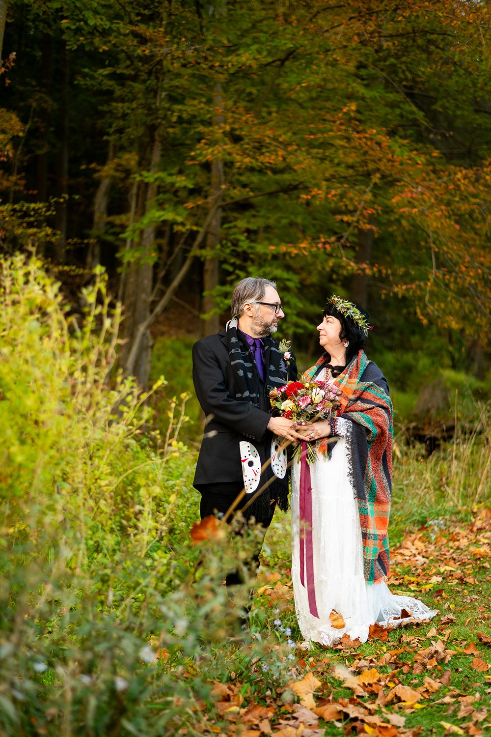 A couple in traditional Celtic-inspired wedding attire stand among autumn leaves in a woodland setting.