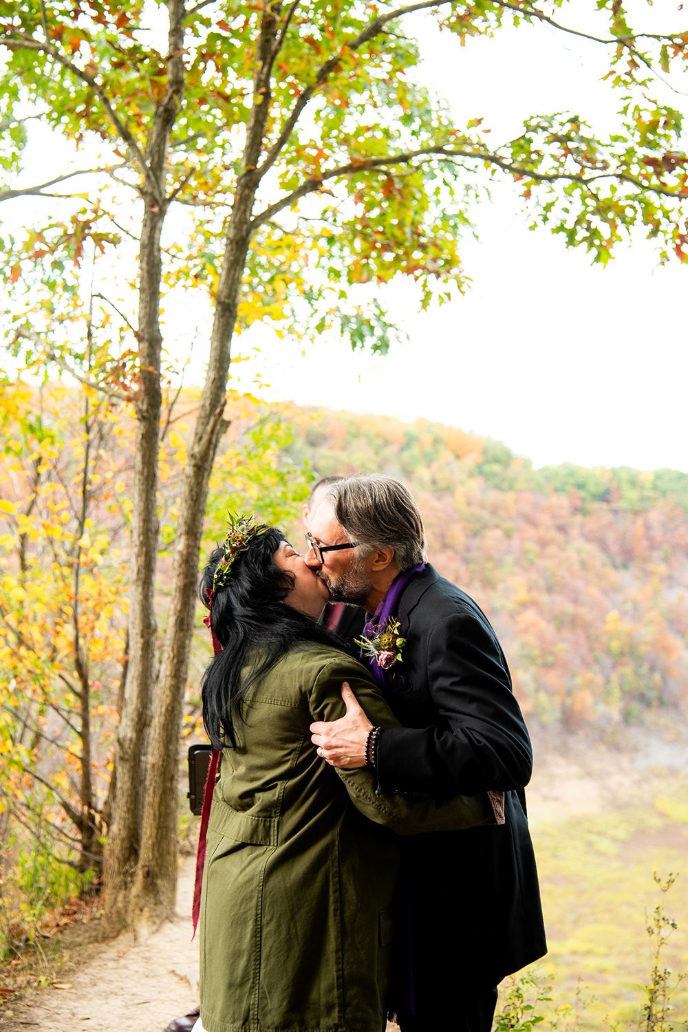 A couple shares their first kiss during an autumn wedding ceremony surrounded by fall colors.
