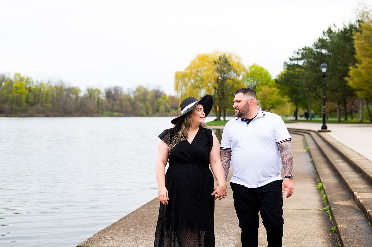 Couple in black and white outfits walking together by a serene lake with stone steps and autumn trees in background.