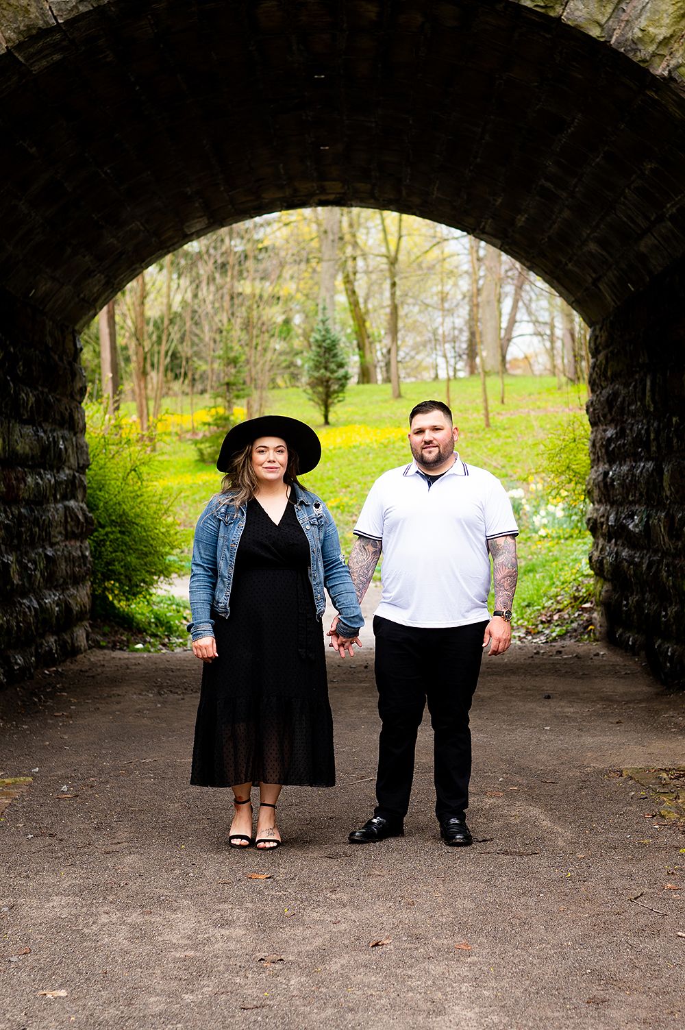 A couple holding hands while standing beneath a stone arch tunnel with greenery visible in the background.