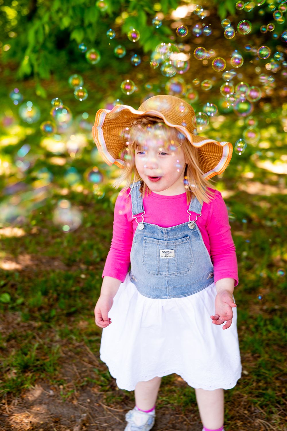 A child wearing overalls, a pink shirt and floppy sun hat plays outdoors surrounded by floating soap bubbles.