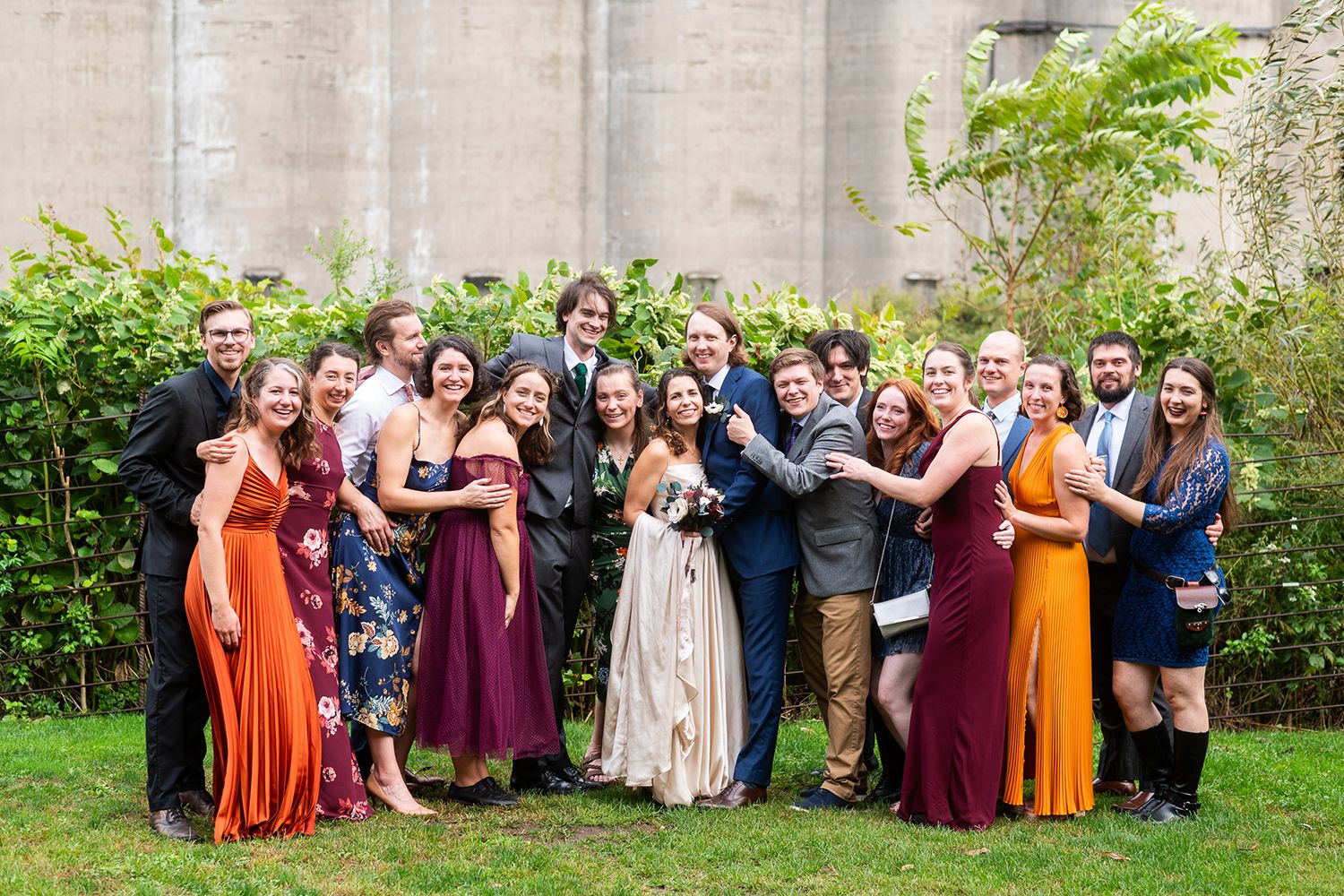 Wedding party celebrates together outdoors wearing mix of burgundy, orange, and navy dresses with greenery backdrop.