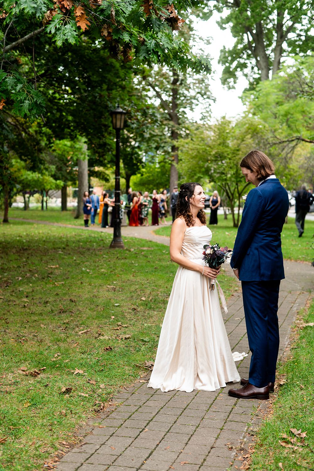 Couple in formal wedding attire sharing a sweet moment on a stone path surrounded by lush green park scenery.