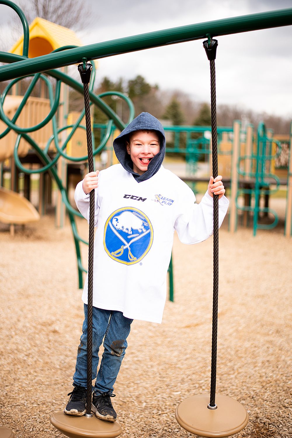 A child wearing a Buffalo Sabres hockey jersey and blue hoodie plays at a playground on the swing set.