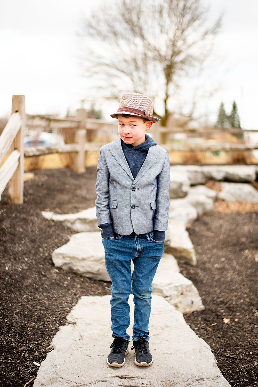 Young child wearing a gray blazer, fedora hat and jeans stands on a stone pathway in an outdoor garden setting.