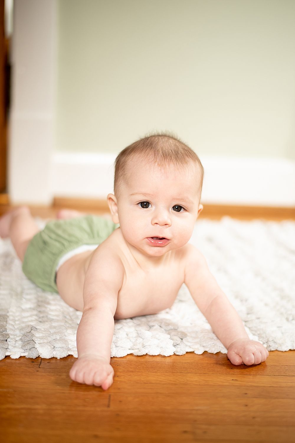A baby in green shorts crawls on hardwood flooring next to white lace fabric.