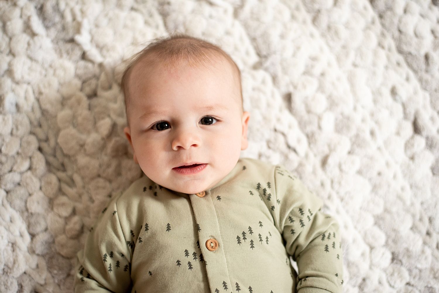 A young infant wearing a sage green cardigan lies on a textured white blanket while looking upward with an expression of joy.