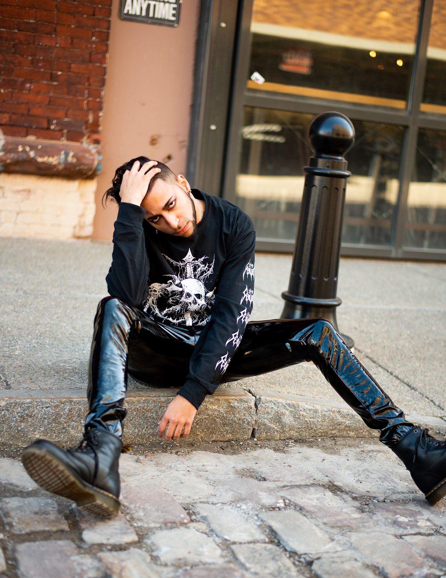 Person in black sweater and boots sits on cobblestone street against brick wall in edgy urban fashion pose.