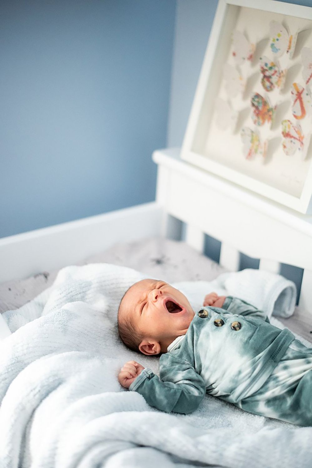 A baby wearing a blue sleeper yawns while lying on white bedding beneath framed nursery artwork.