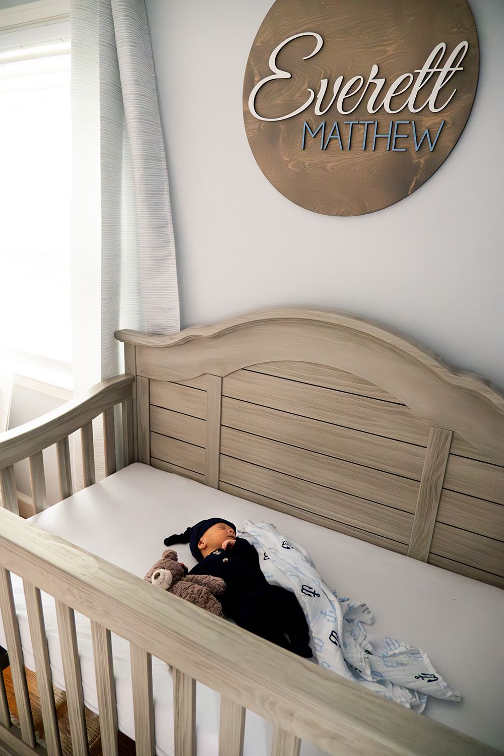 A wooden crib with gray finish and round name sign reading 'Everett Matthew' mounted on blue wall above.
