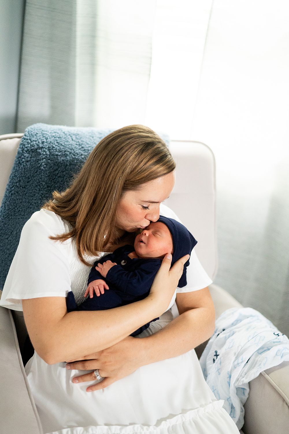 A tender moment between a parent and newborn baby while sitting in a cozy white armchair near a window.