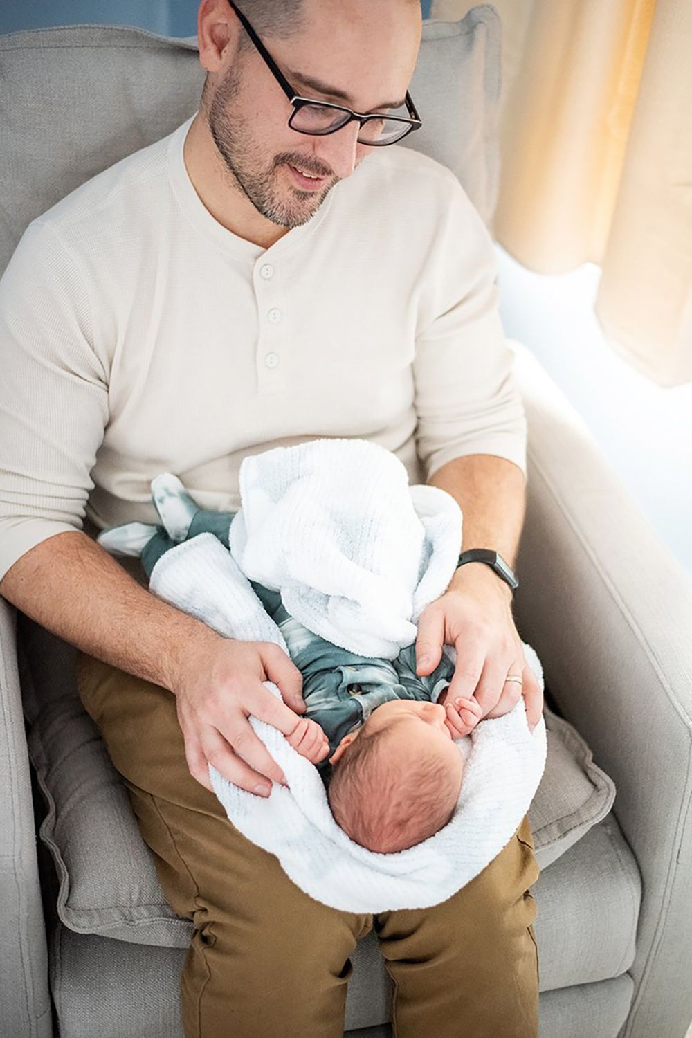 A gentle moment as a person in glasses cradles a swaddled infant on a grey armchair.