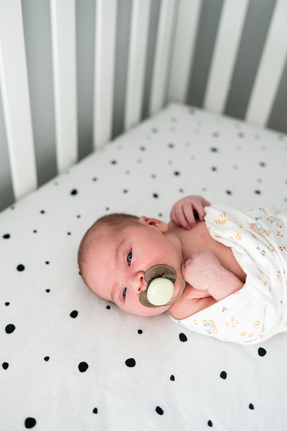 Sleeping newborn in polka dot crib bedding wearing white sleepwear with pacifier.