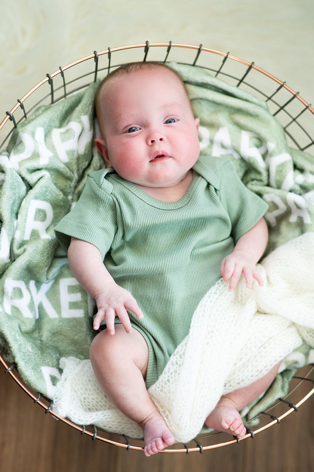 Baby wearing mint green onesie lies in round basket with patterned blanket and white knit fabric.
