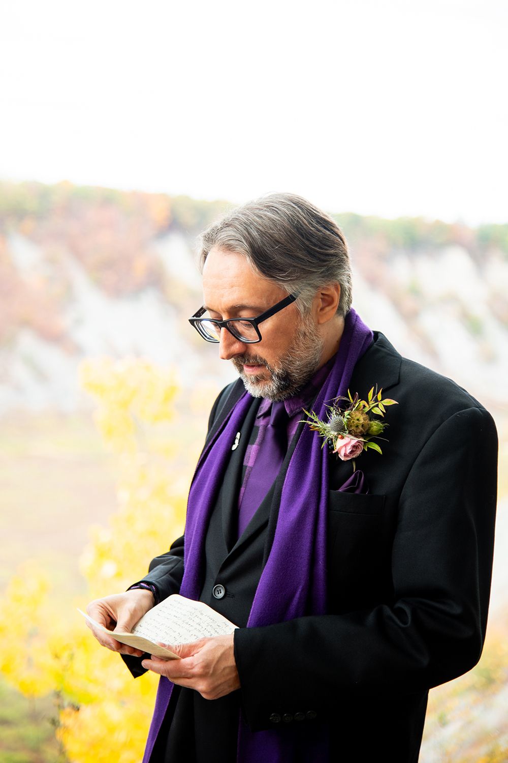 Someone wearing glasses, a black suit and purple scarf reads from papers with yellow autumn foliage in background.