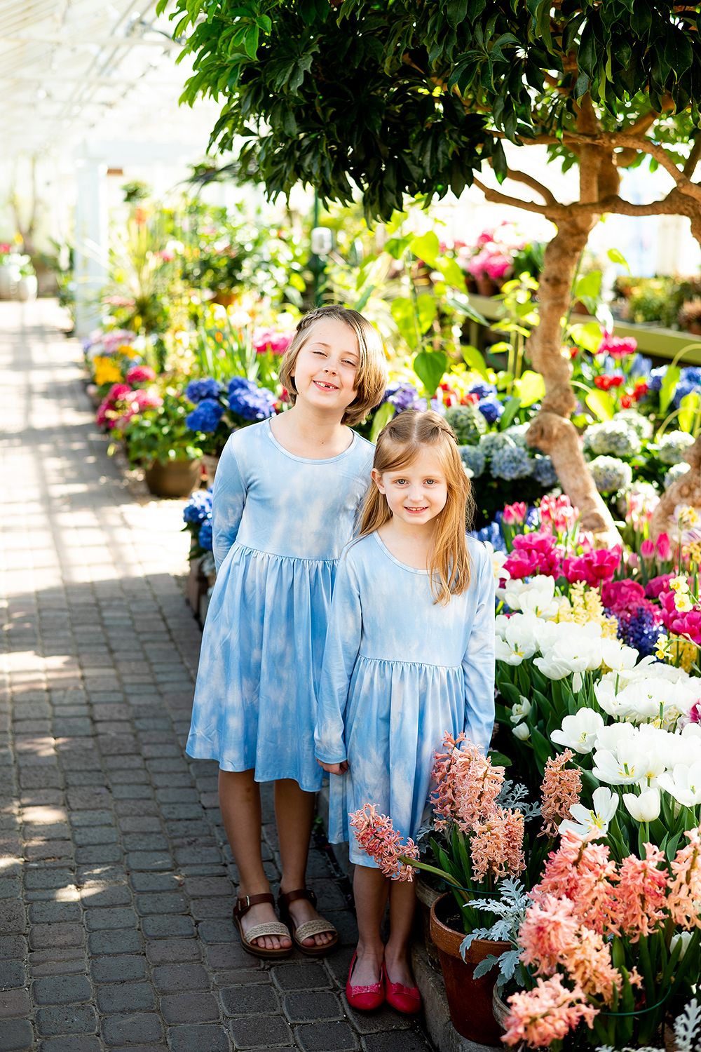 Two children in matching blue dresses stand together among vibrant spring flowers in a garden pathway.