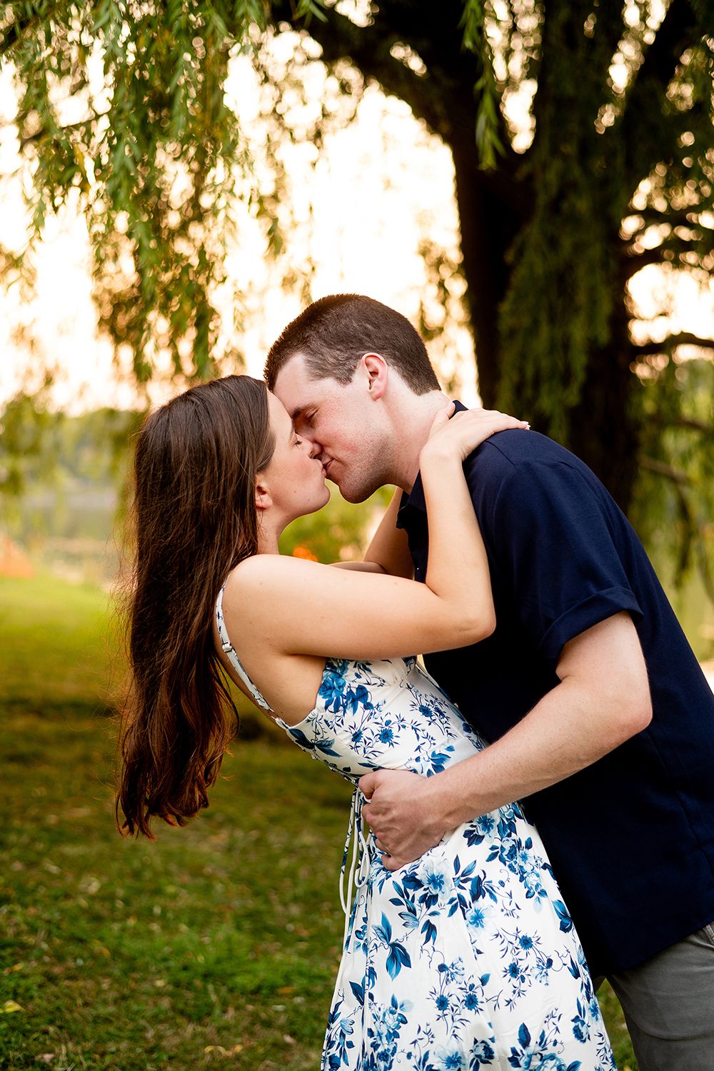 Romantic couple embraces and kisses under willow trees at sunset in a park setting.