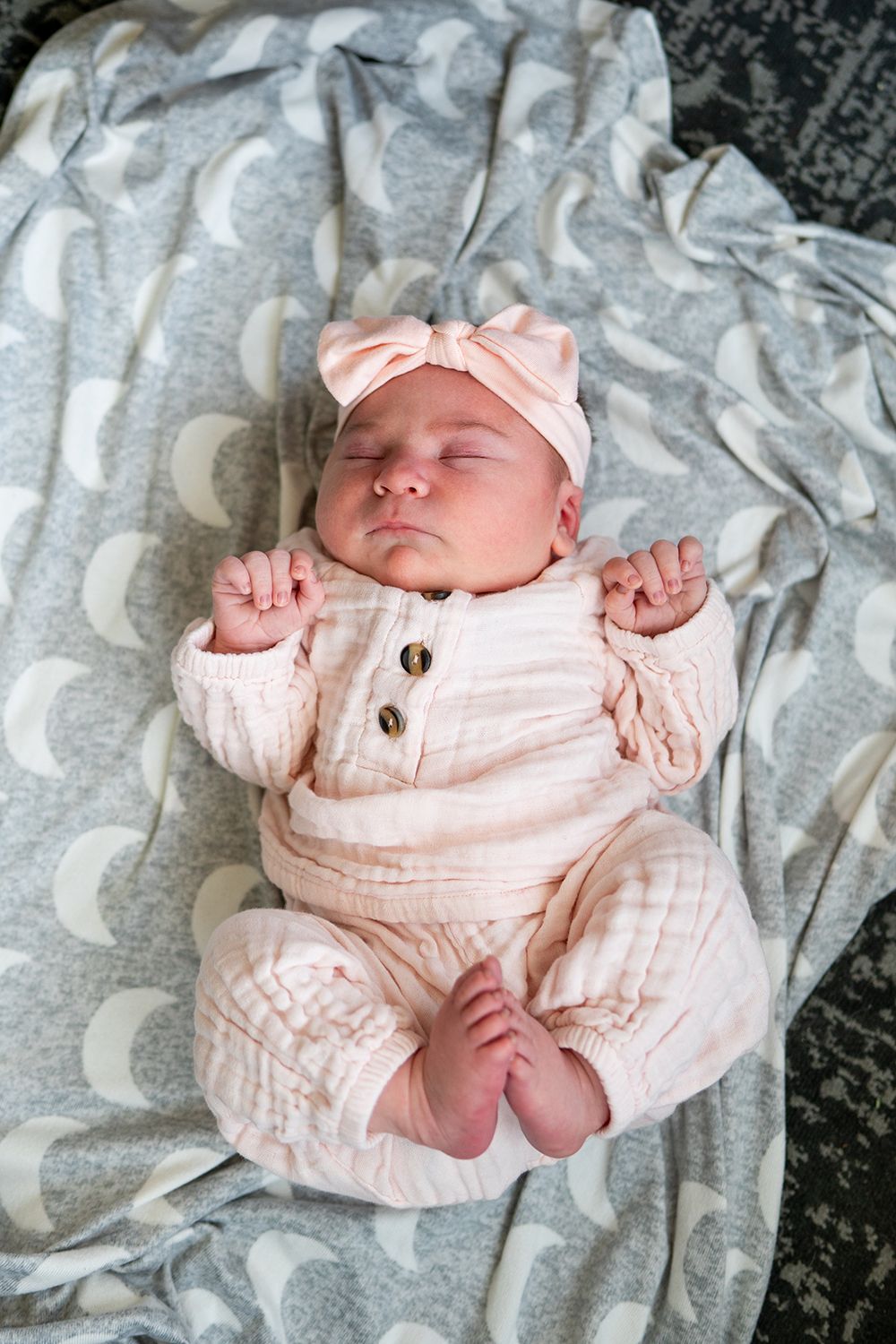 Sleeping newborn wearing soft pink romper with bow headband rests on gray patterned blanket.