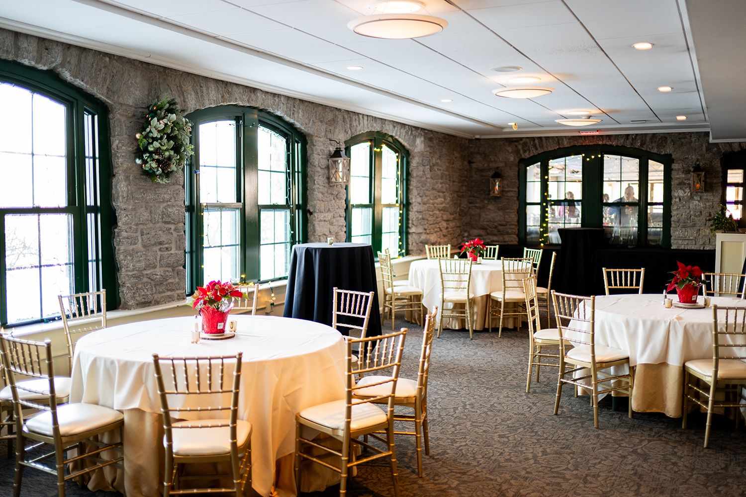 Elegant banquet hall with brick walls, arched windows, round tables with white tablecloths, and gold chiavari chairs.