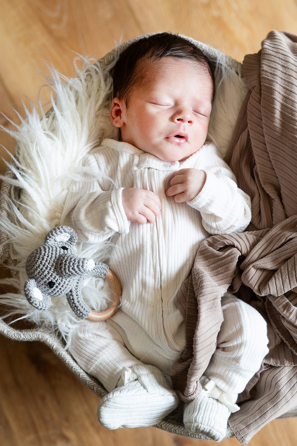 Sleeping newborn in white knit outfit rests on fuzzy cream blanket with gray knit toy nearby.