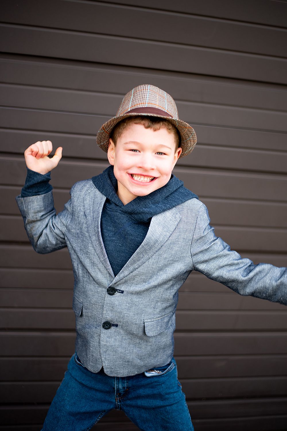 Energetic pose in gray blazer and fedora hat against dark wall showing playful movement and happiness.