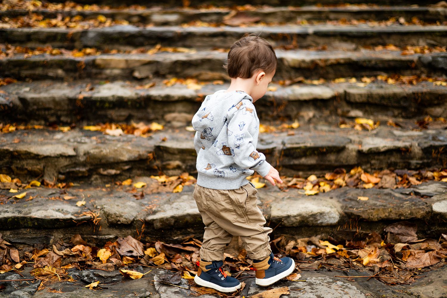 Small child in blue sneakers and beige pants exploring stone steps strewn with fall foliage.