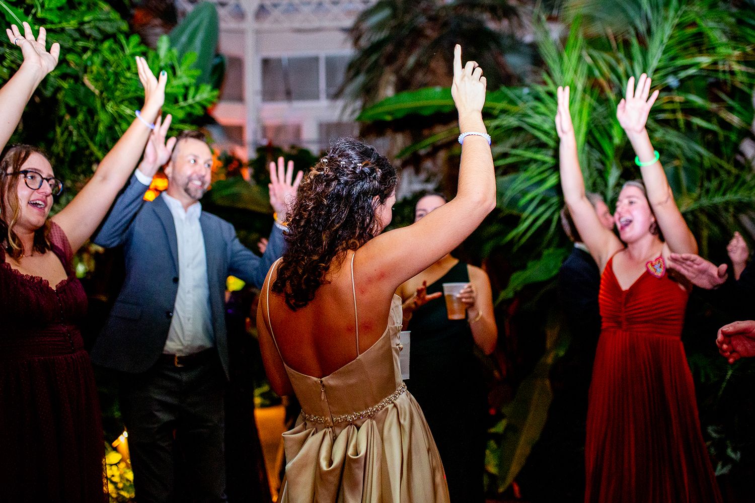 People dancing with hands raised in celebration at an upscale party event surrounded by tropical plants and decor.