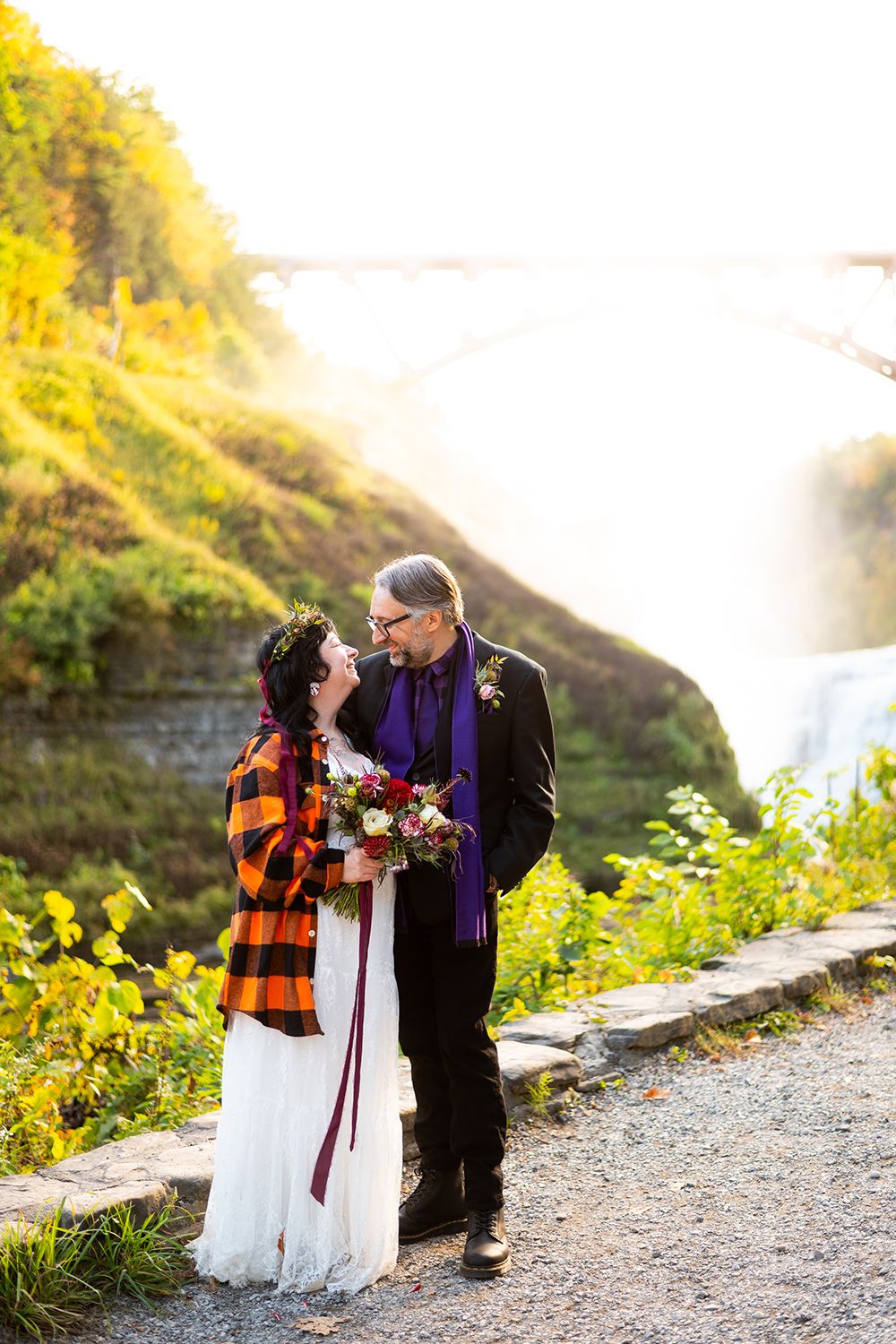 A couple shares an intimate autumn moment on a sunlit gravel path surrounded by yellow wildflowers.
