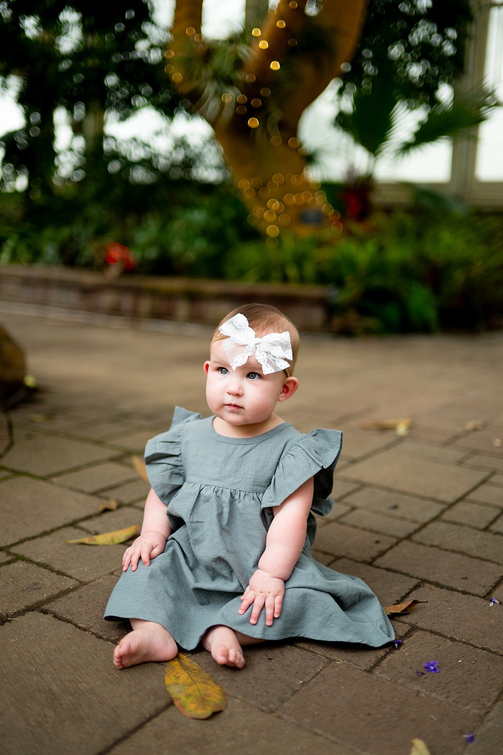 Small child in gray dress sits on stone pavers during outdoor photoshoot.