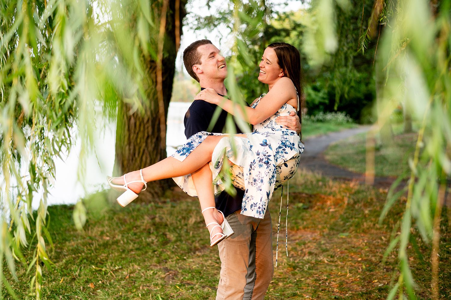 A romantic couple shares a playful moment under drooping willow tree branches in a sunlit park setting.