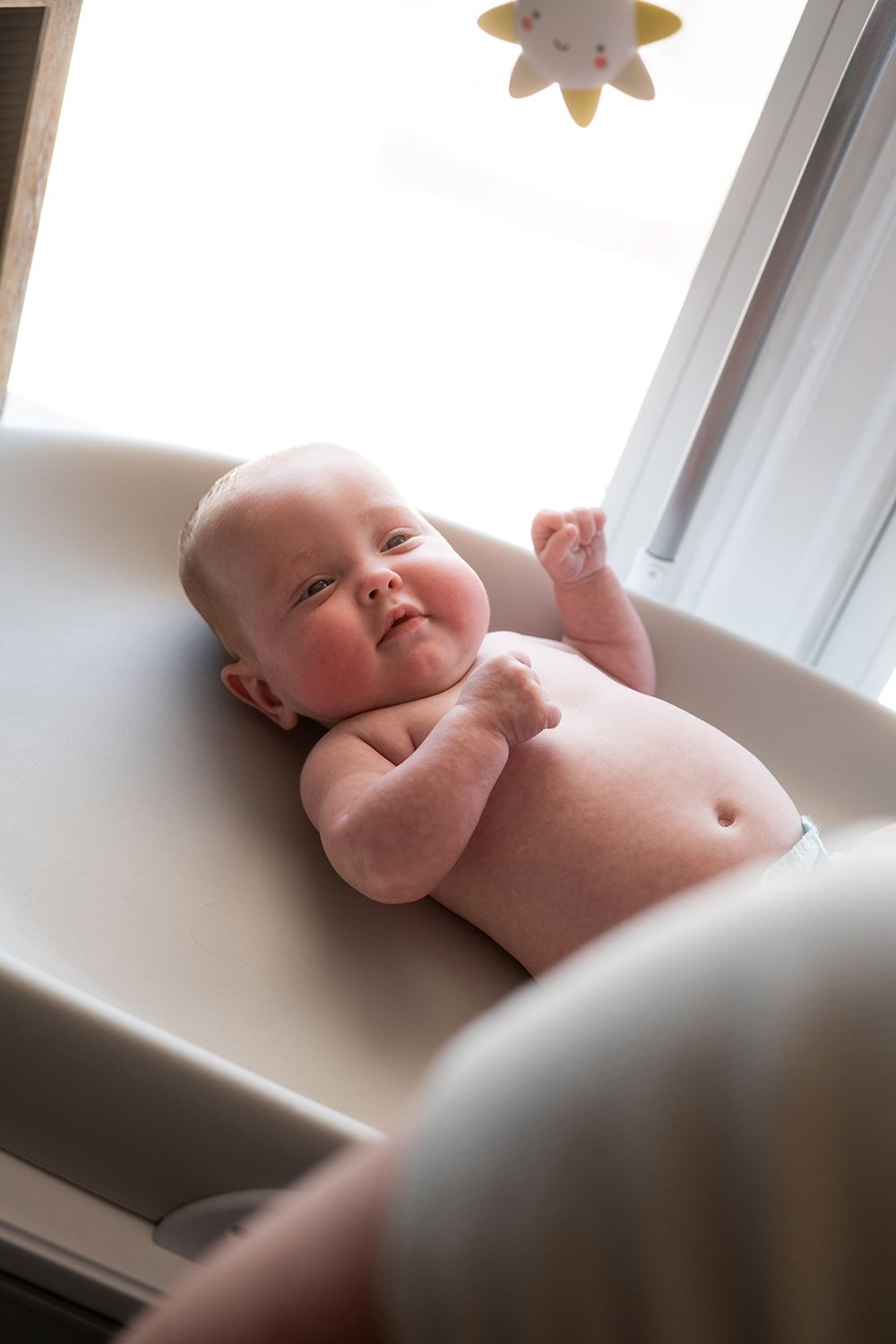 Baby lying on back under window with star decoration overhead makes expressive face at camera.