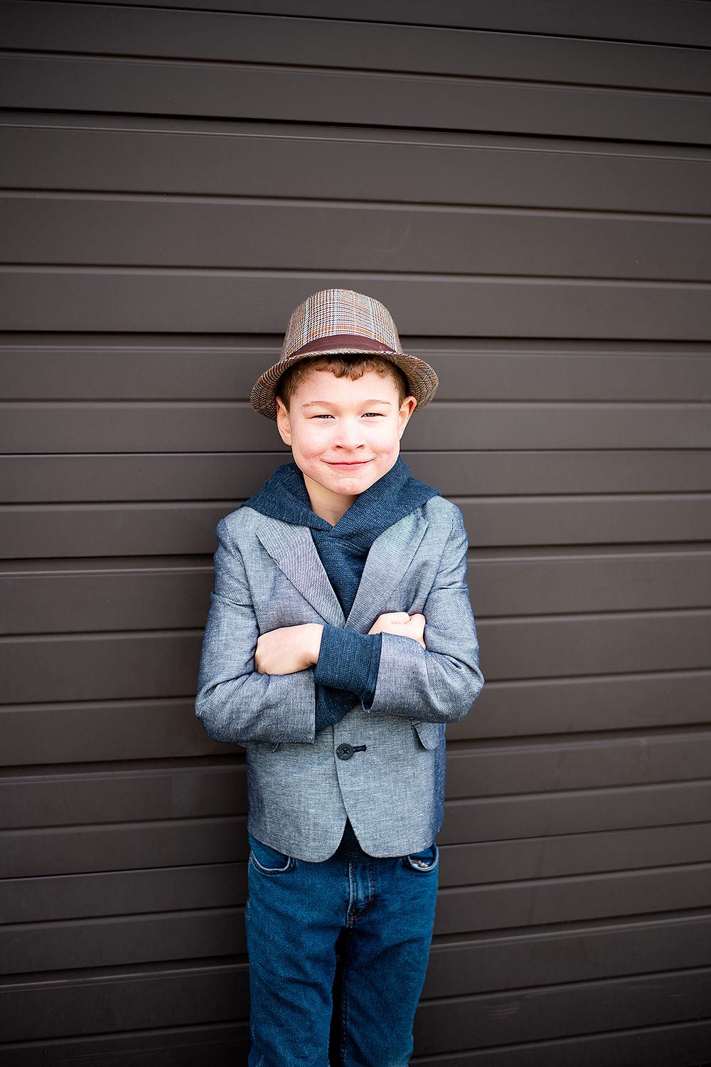 Young person in a gray blazer, blue shirt and fedora hat poses with arms crossed against dark wall.