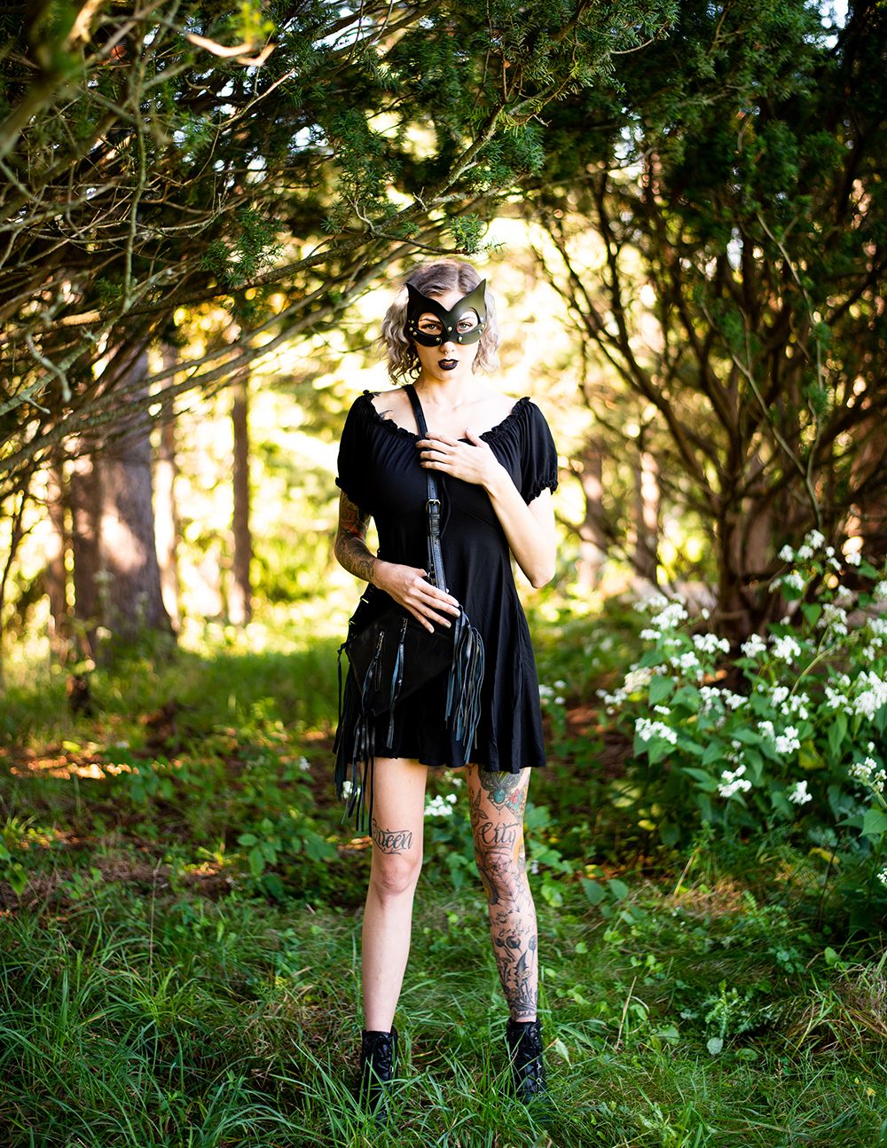 A person in a black velvet dress and cat mask poses in a sunlit forest clearing near white wildflowers.